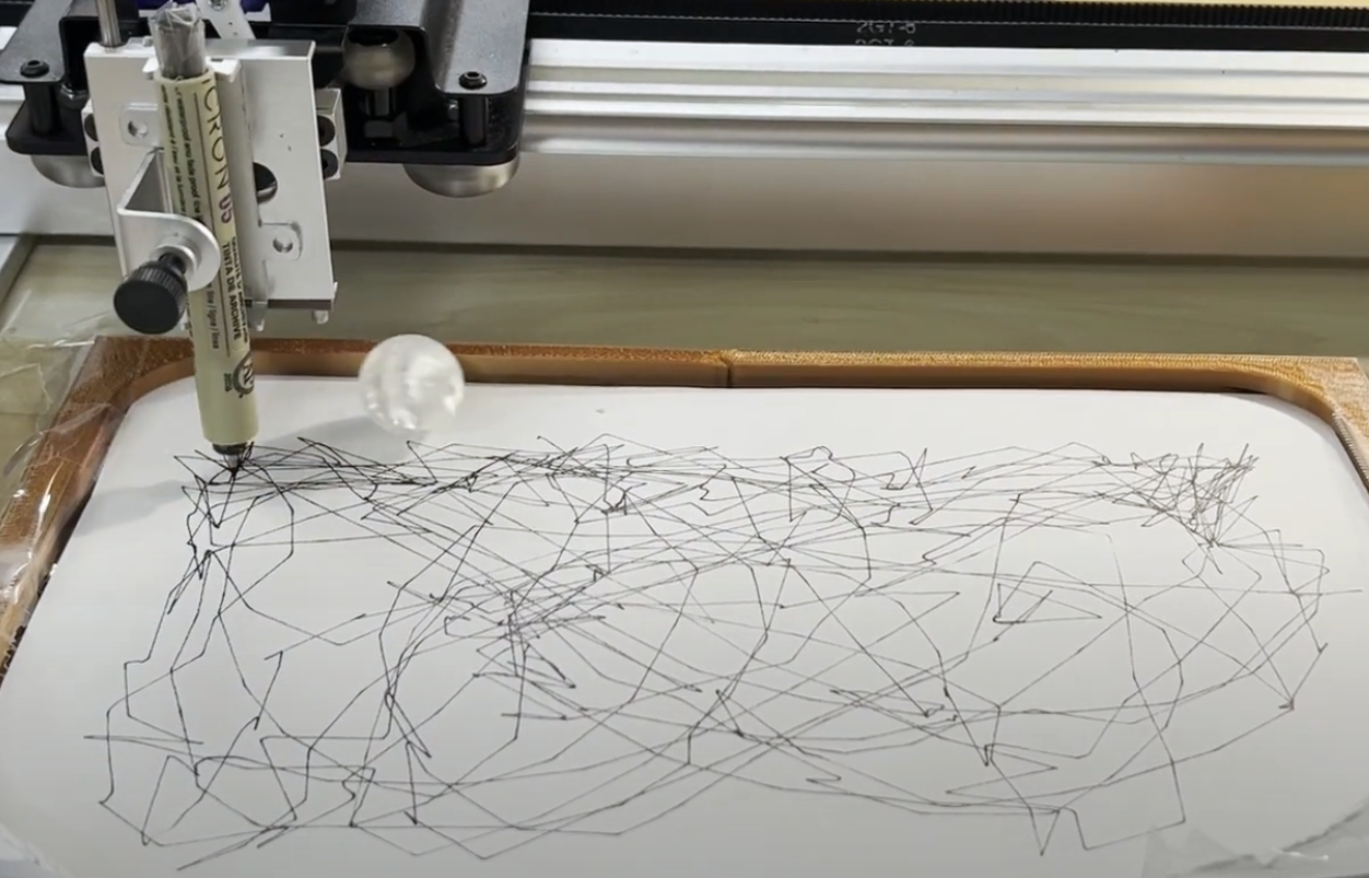 The AxiDraw mid-run: a Micron pen in the carriage, a glass marble on the paper beside it, and a dense tangle of plotted lines inside the printed arena