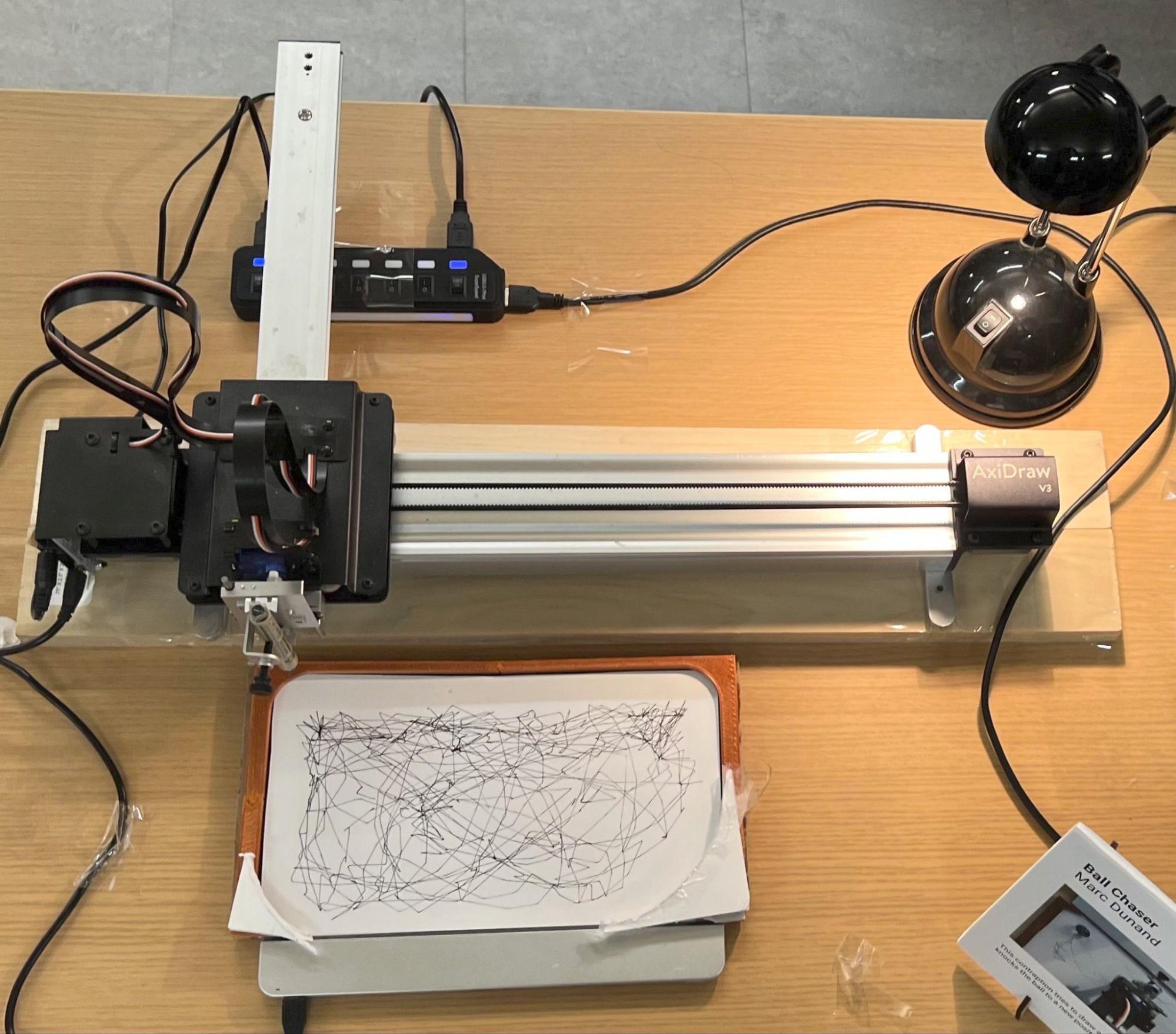 The installed piece on a table: the AxiDraw on a wooden base, the printed arena holding paper over the Sensel pad, a desk lamp and a printed placard - and no camera anywhere