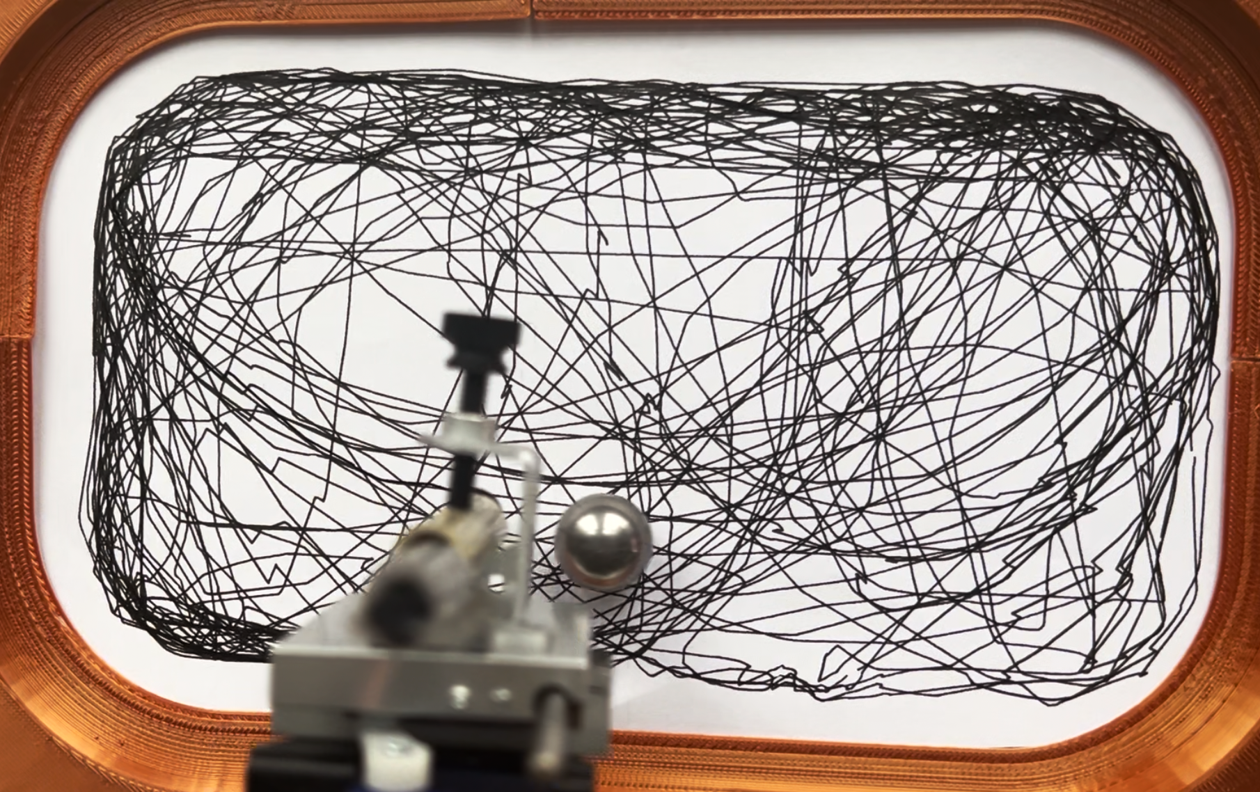 Overhead view inside the orange arena: a dense finished drawing, the pen carriage, and the metal sphere resting just beside the nib